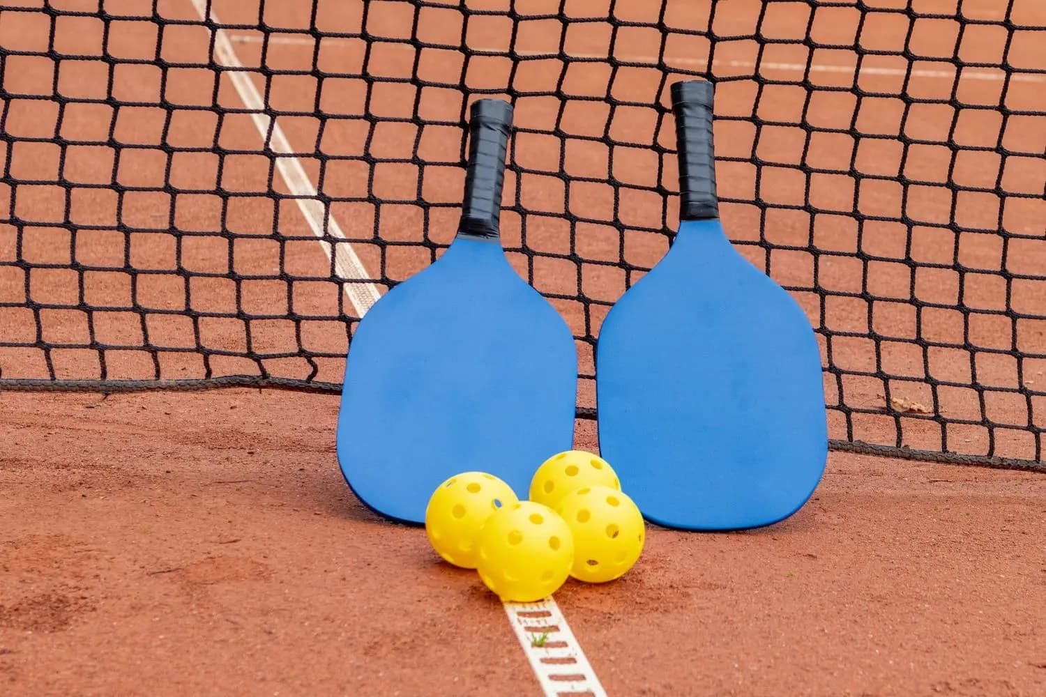 Two pickleball paddles and balls by the net on a clay court